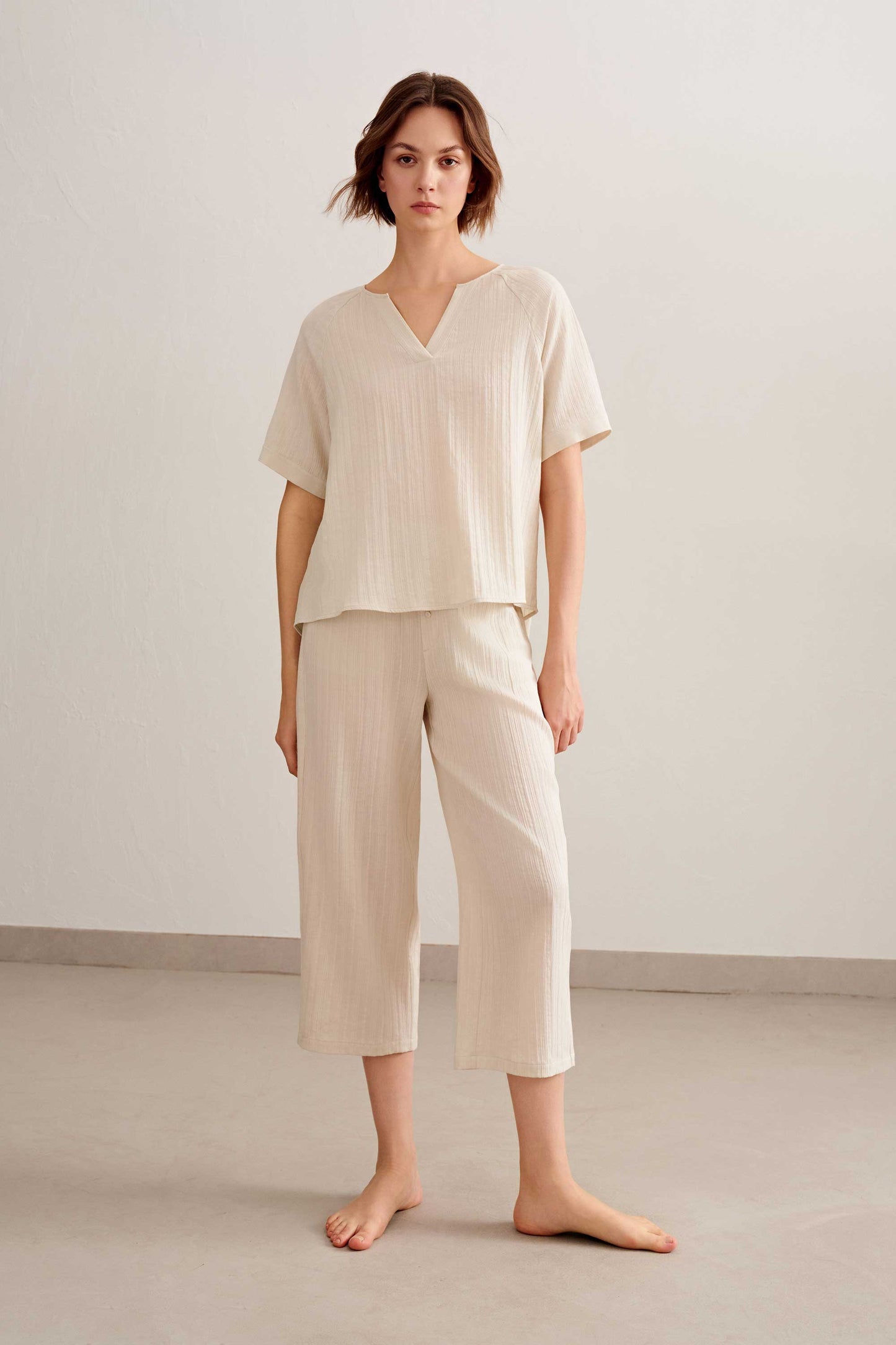 a woman standing facing the front, wearing a light beige short sleeve v neckline tee with light beige cropped pants.