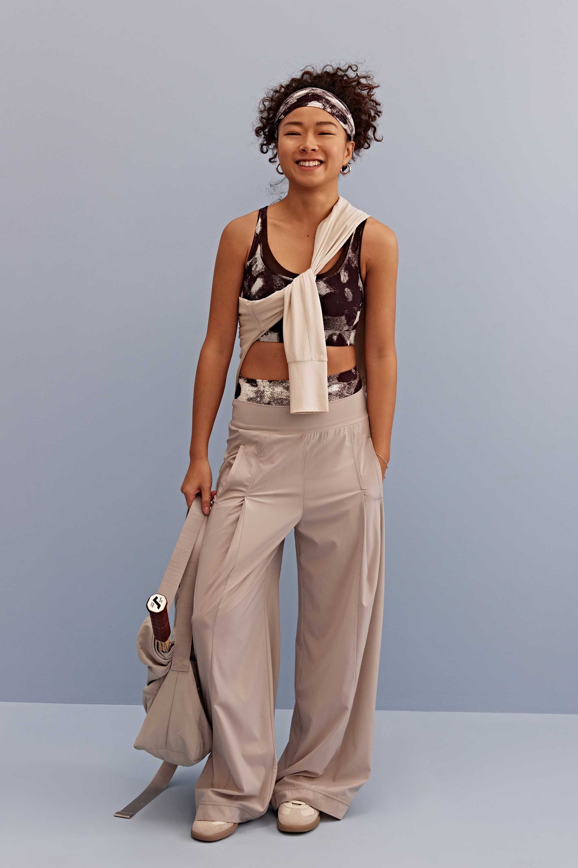 a woman wearing a metallic sports bra and headband, pair with a light beige pants and shoes. holding a light beige bag
