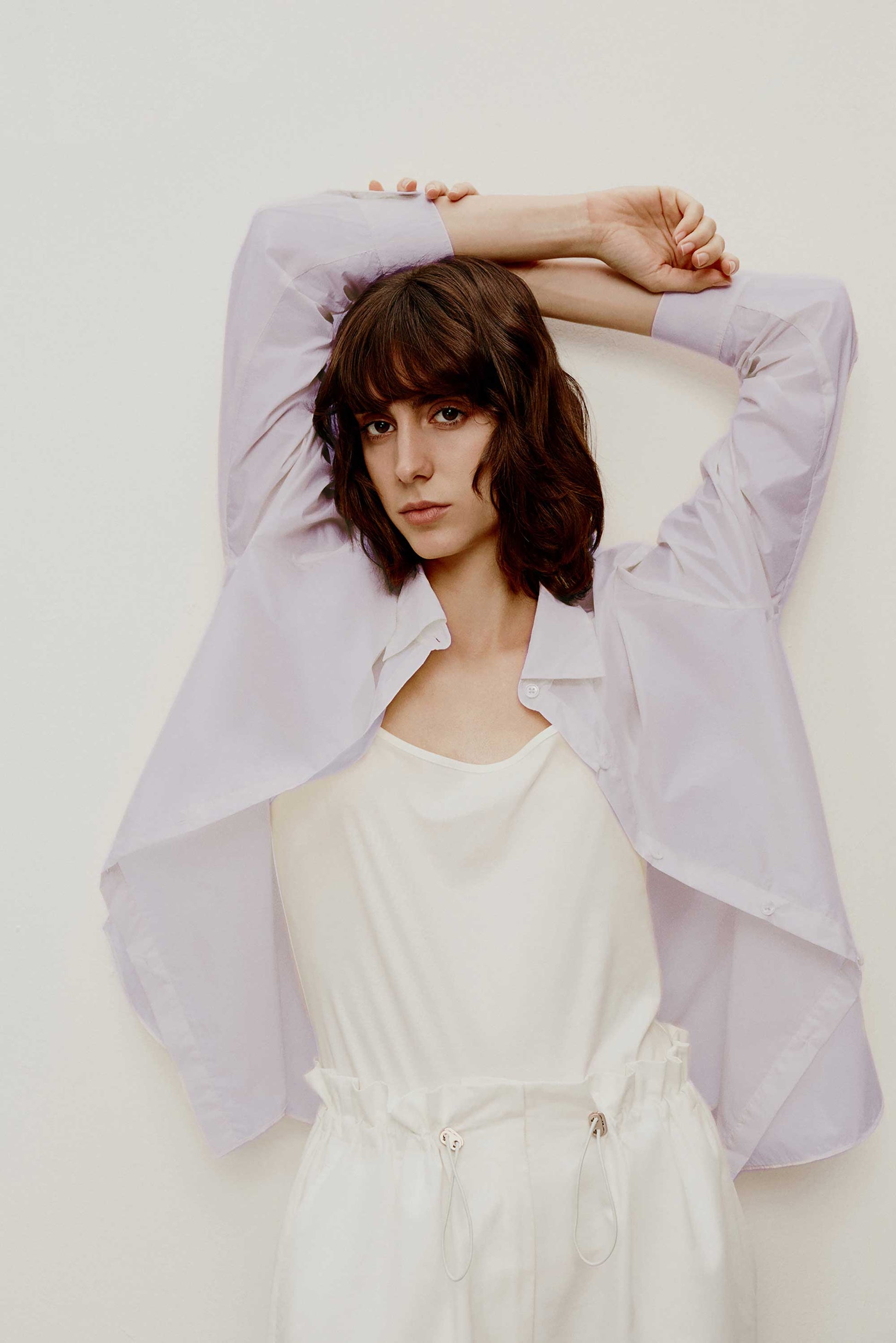 a woman lifts her arms with short brown curly hair wearing a light purple shirt. a white camisole under the shirt and a white pants