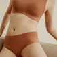 A person wears a seamless, brown Barely Zero Classic Bra + Brief Set and matching Mid-Waist Brief in a beige room. The focus is on the comfortable, form-fitting underwear designed for a smooth silhouette.