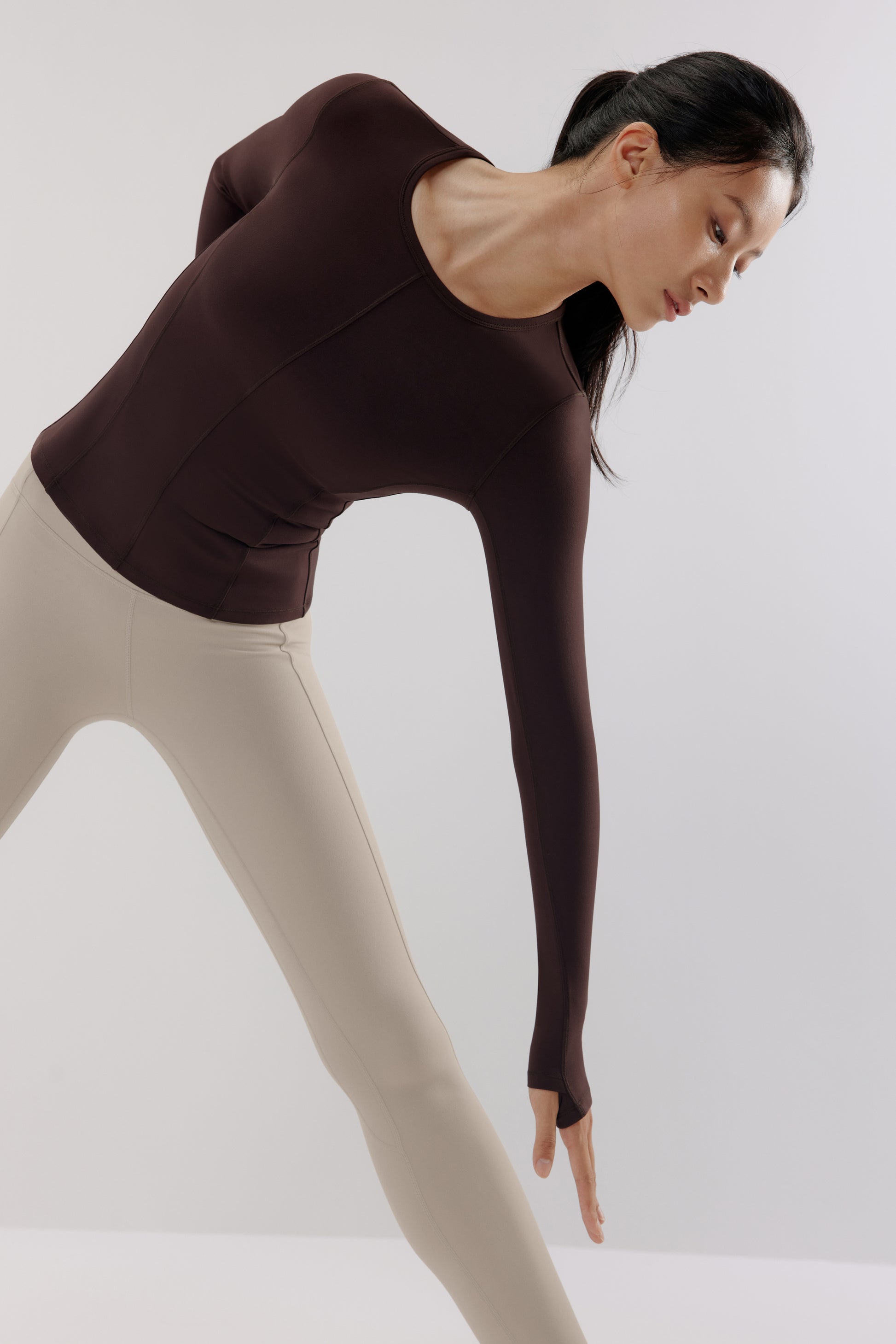A woman wears a brown mousse long sleeve sports top and grey leggings and doing Yoga.