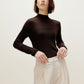 A woman wears a brown silky wool mock neck sweater and white pants.