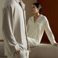 on the front, a man wearing a beige pajama set standing. on the back, a woman wearing a cream pajama set sitting on a white sofa