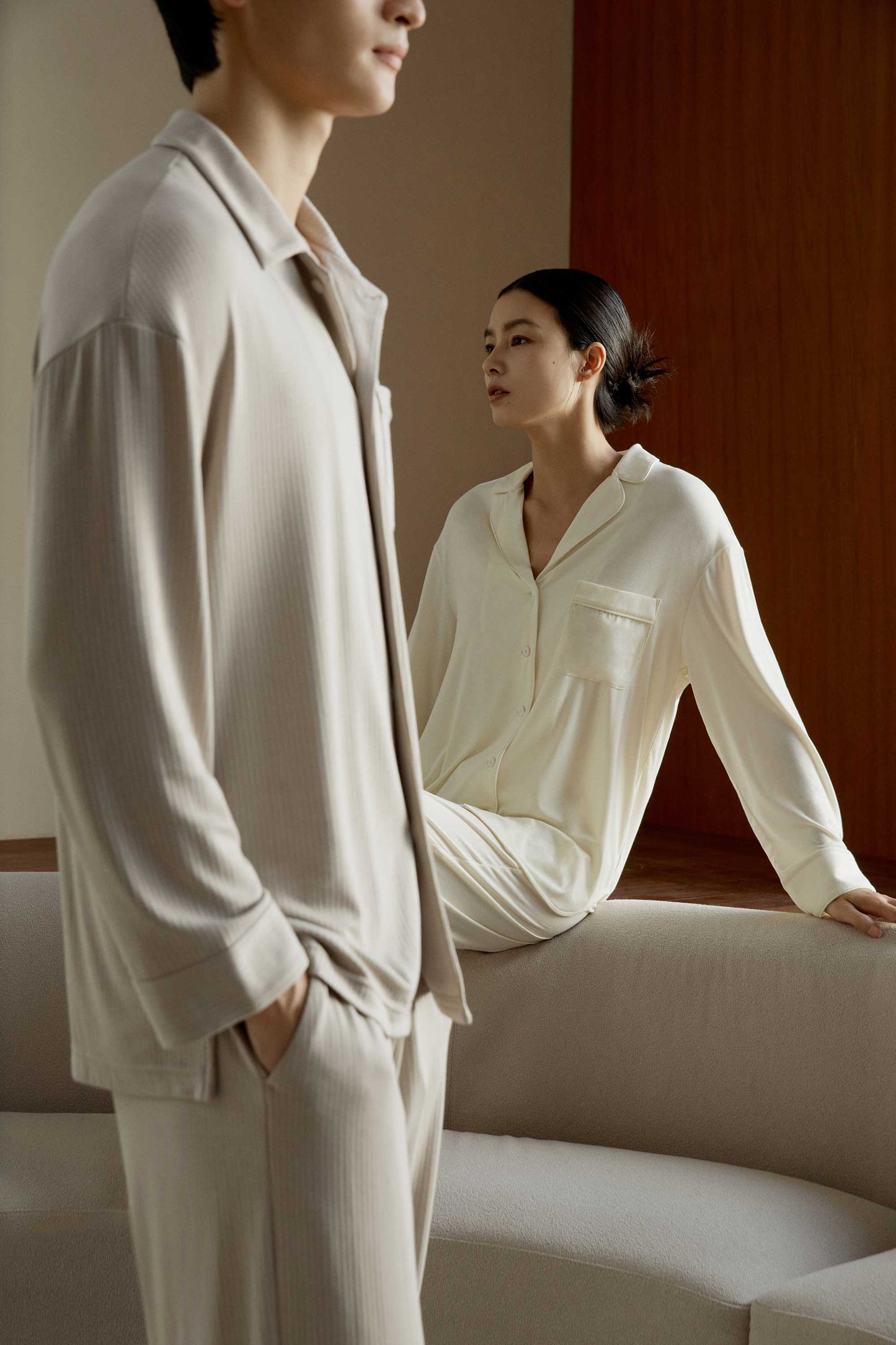 on the front, a man wearing a beige pajama set standing. on the back, a woman wearing a cream pajama set sitting on a white sofa