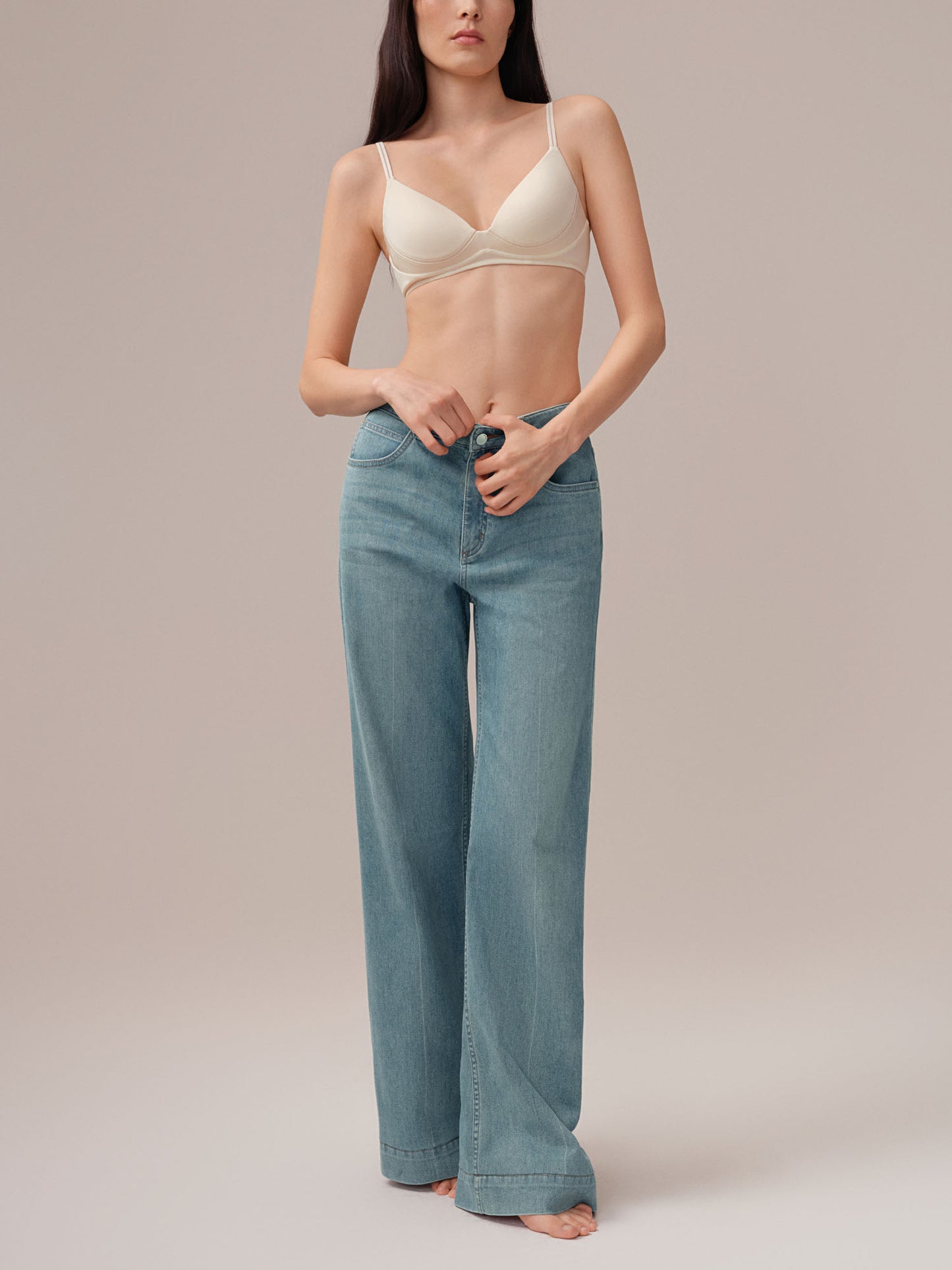 Model wearing a beige bra and blue jeans against a plain background