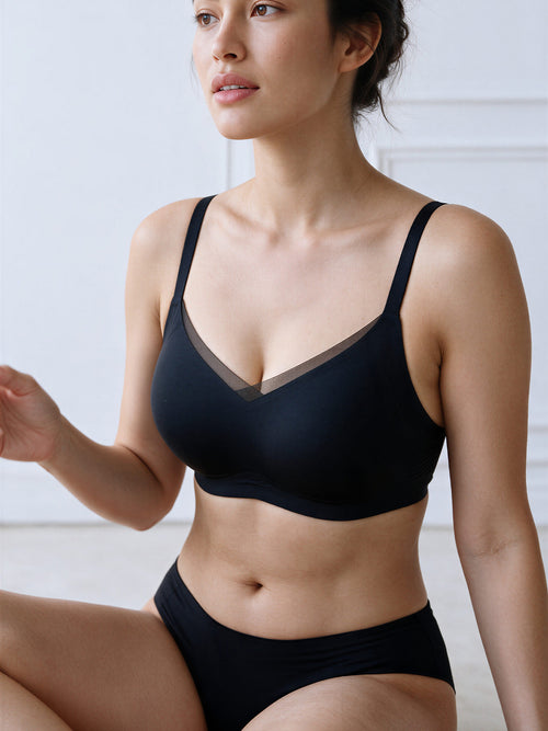 A woman wearing a black wireless bra and matching black underwear, posed in a bright, minimalist indoor setting.