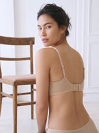 A woman wearing a beige wireless bra and matching underwear, photographed from the back in a bright indoor setting with a wooden chair visible on the left. Toasted Almond