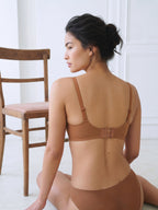A woman wearing a matching tan bra and underwear set, shown from the back in a brightly lit indoor setting with a wooden chair to the left. Sandstone