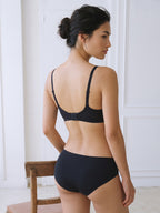 A woman shown from the back wearing a black bralette and matching high-waisted briefs in a bright indoor room with white paneled walls and a wooden chair partially visible. Black