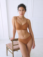 A woman modeling a matching two-piece bikini set in a warm terracotta brown color, standing next to a wooden chair in a bright, white-paneled room. Sandstone