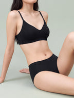 A woman wearing a black bralette and matching high-waisted briefs, posed against a solid light gray studio background.black