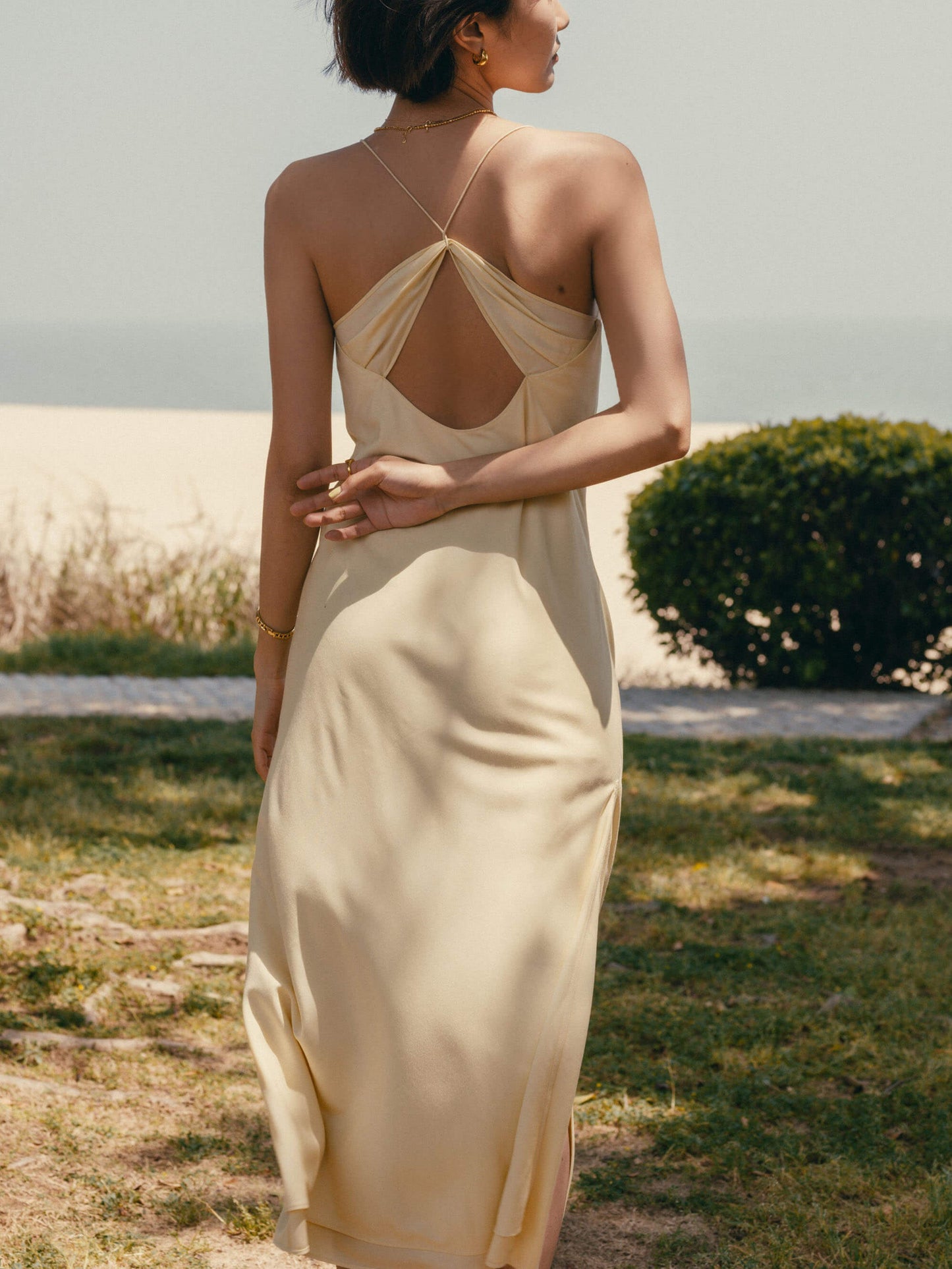 Back view of woman standing on grass wearing cream colored dress with thin straps and cross back design