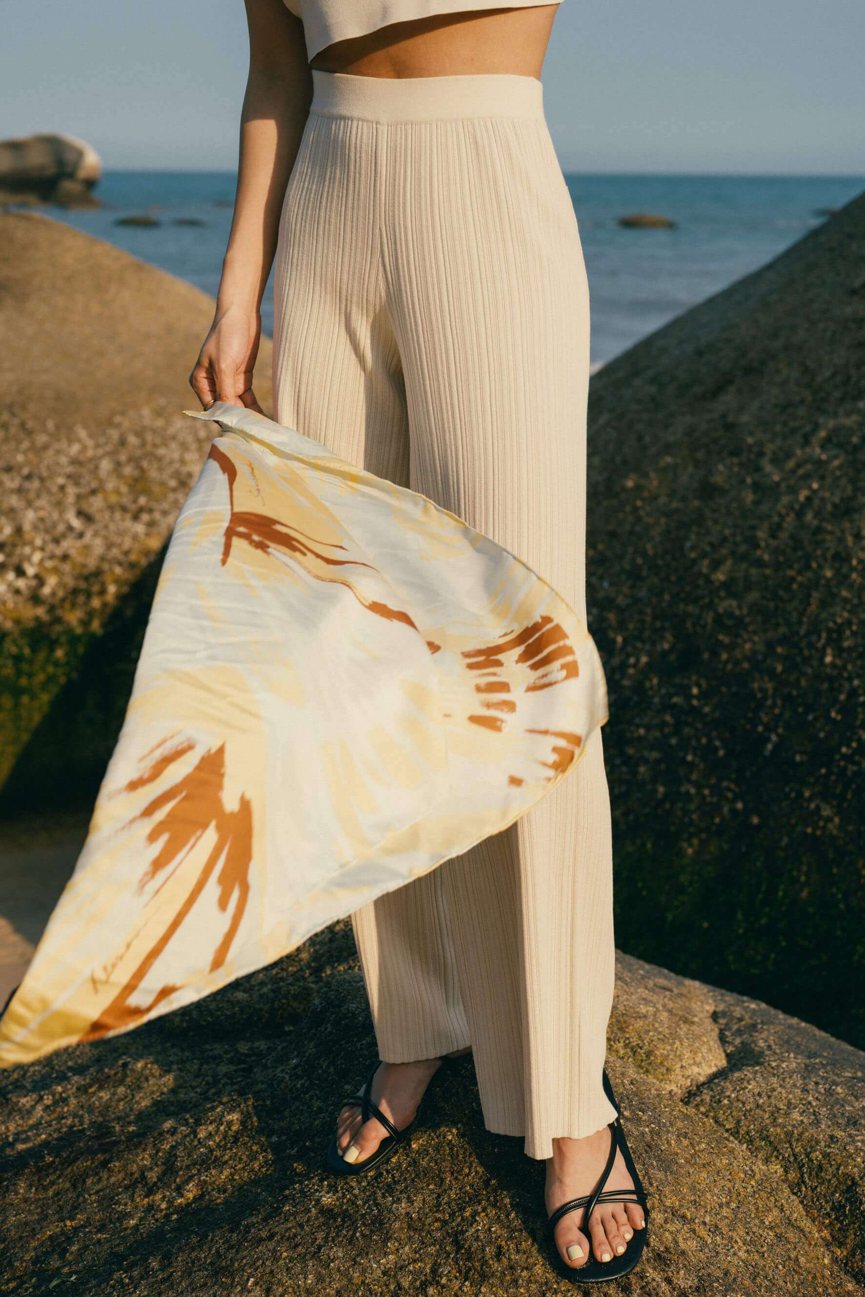 Woman standing on rock wearing off white knit pants holding printed scarf blowing in the wind