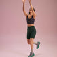 a woman throwing the ball wearing a black long sports bra and green biker shorts. pair with grey socks and green shoes.