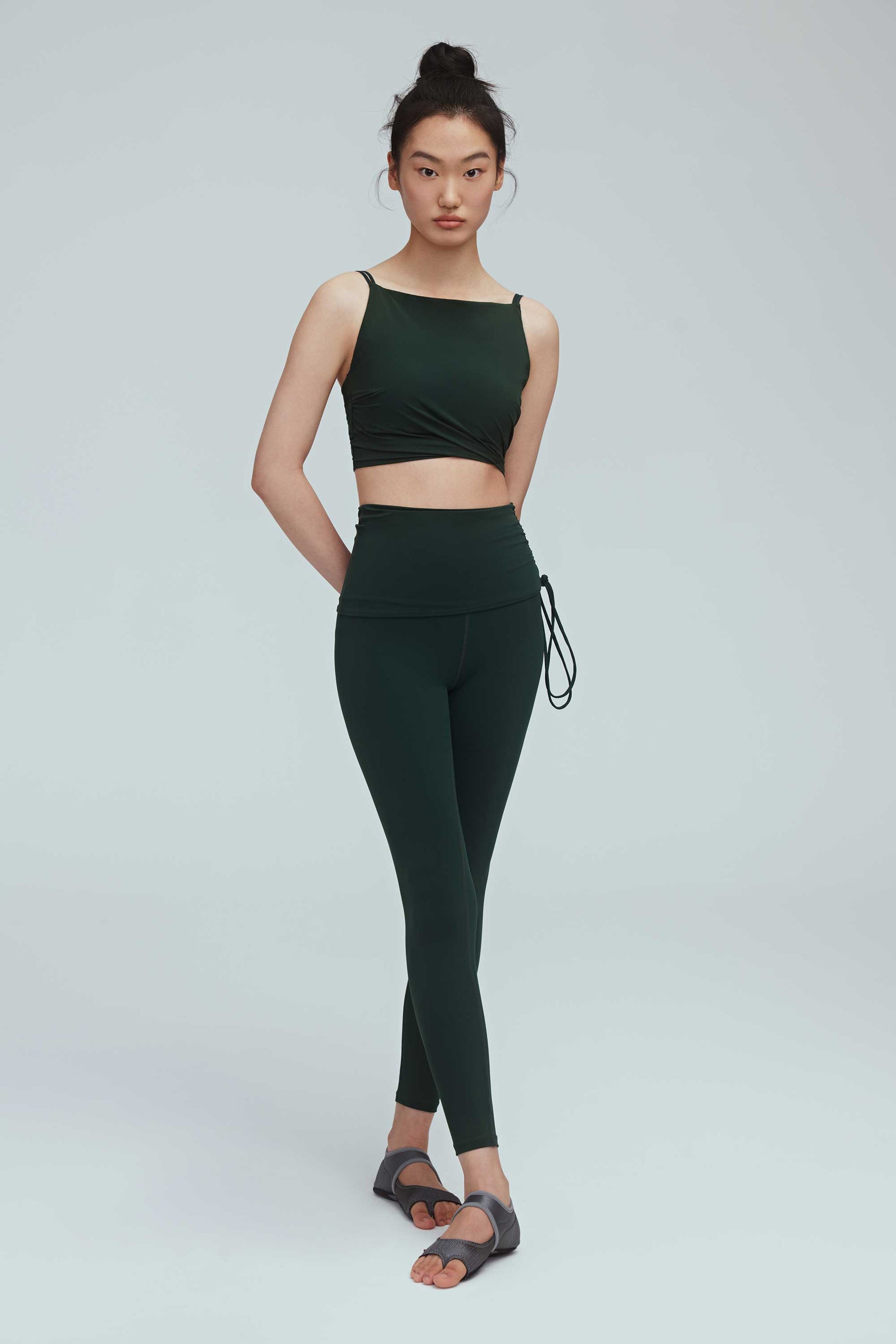a woman wearing a crew neck dark green sports bra with pleats details and a pair of matching leggings with double layered waist and side knot.