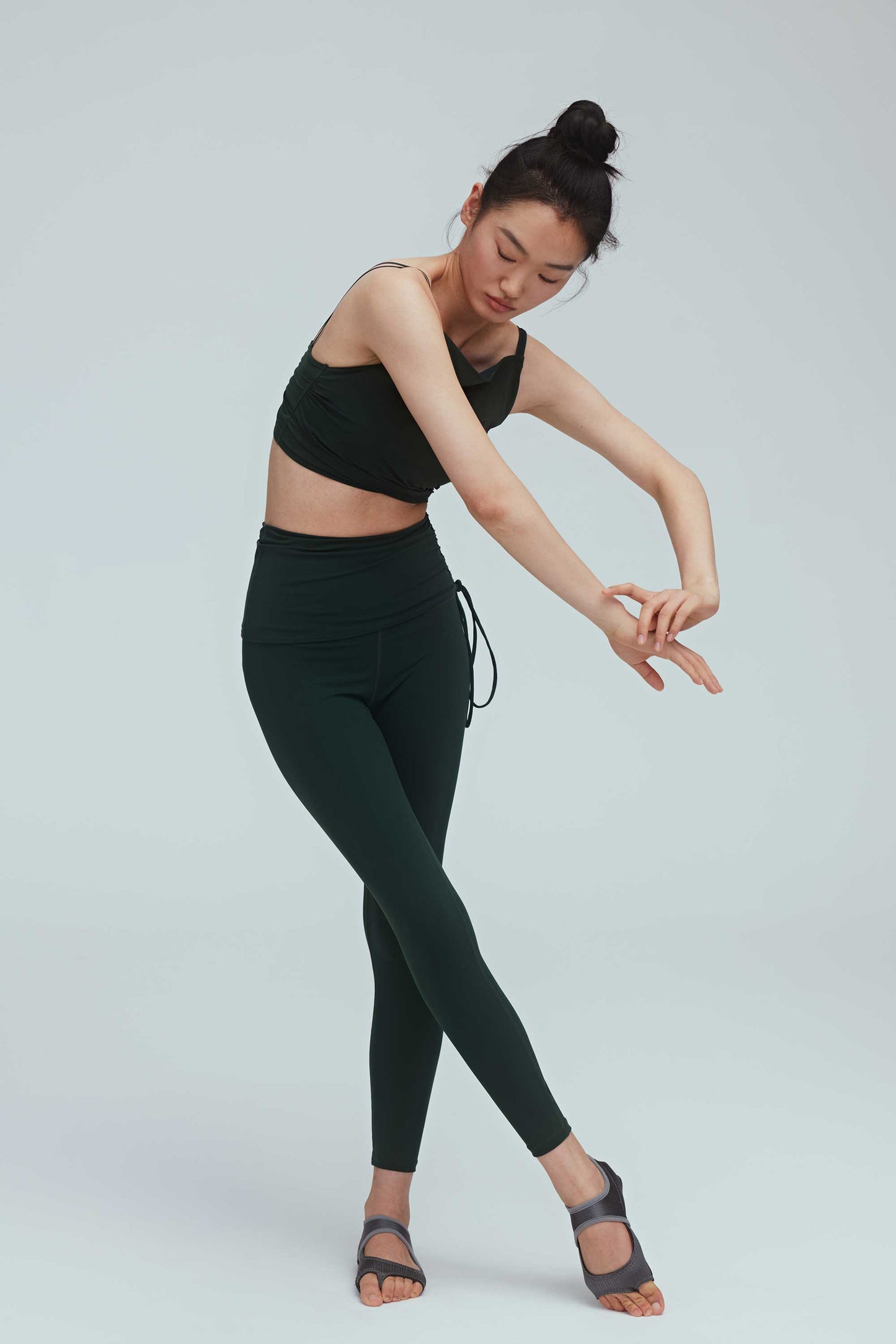a woman doing a dance pose wearing a crew neck dark green sports bra with pleats details and a pair of matching leggings with double layered waist and side knot.