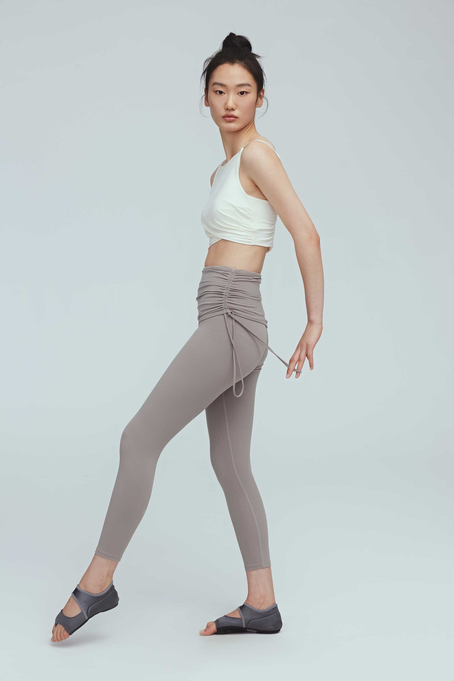 full body side view of a woman wearing a crew neck white sports bra with pleats details and a pair of grey leggings with double layered waist and side knots.