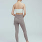 full body back view of a woman wearing a white double strap sports bra with pleats details and a pair of grey leggings with double layered waist and side knots.