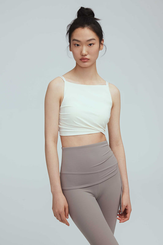 a woman wearing a crew neck white sports bra with pleats details and a pair of grey leggings with double layered waist.