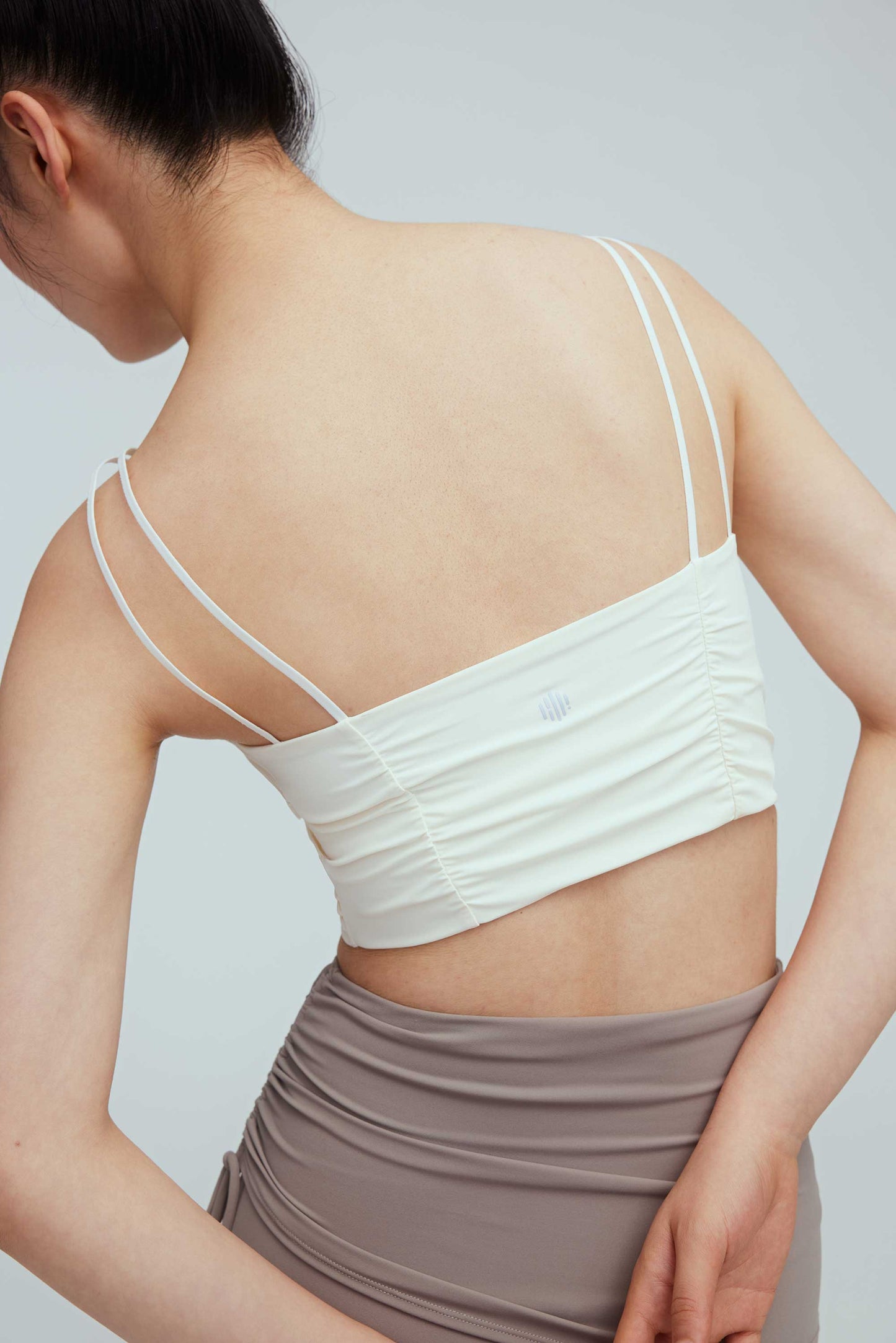 the back of a woman wearing a white sports bra with pleats details and a pair of grey leggings with double layered waist.