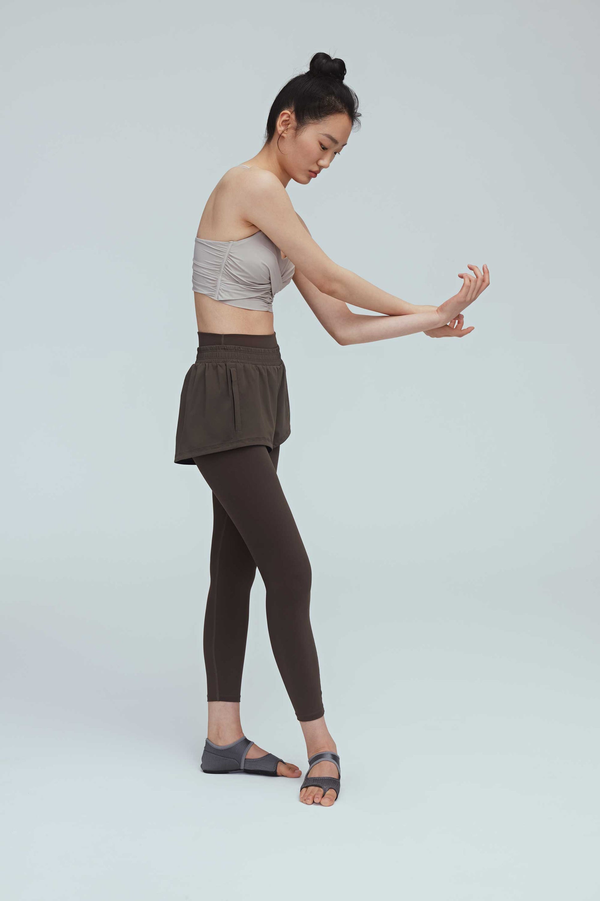 full body side view of a woman with dance pose wearing a light grey color v-neck sports bra with ruched details and a pair of black leggings layered with black shorts