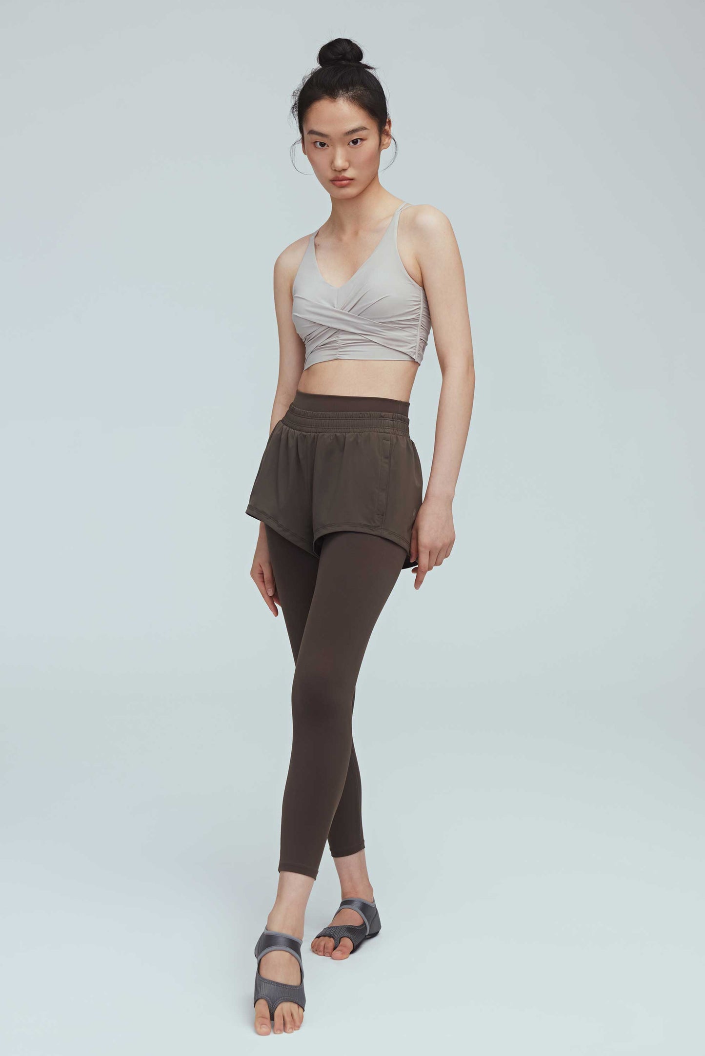 full body front view of a woman wearing a light grey color v-neck sports bra with ruched details and a pair of black leggings layered with black shorts