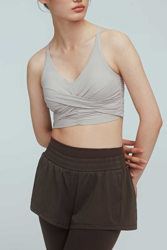 a woman wearing a light grey color v-neck sports bra with ruched details and  a pair of black leggings layered with black shorts 