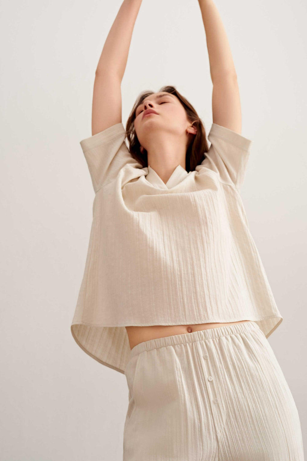 a woman lifts her arms up wearing a a woman wearing a light beige short sleeve tee and pants. 