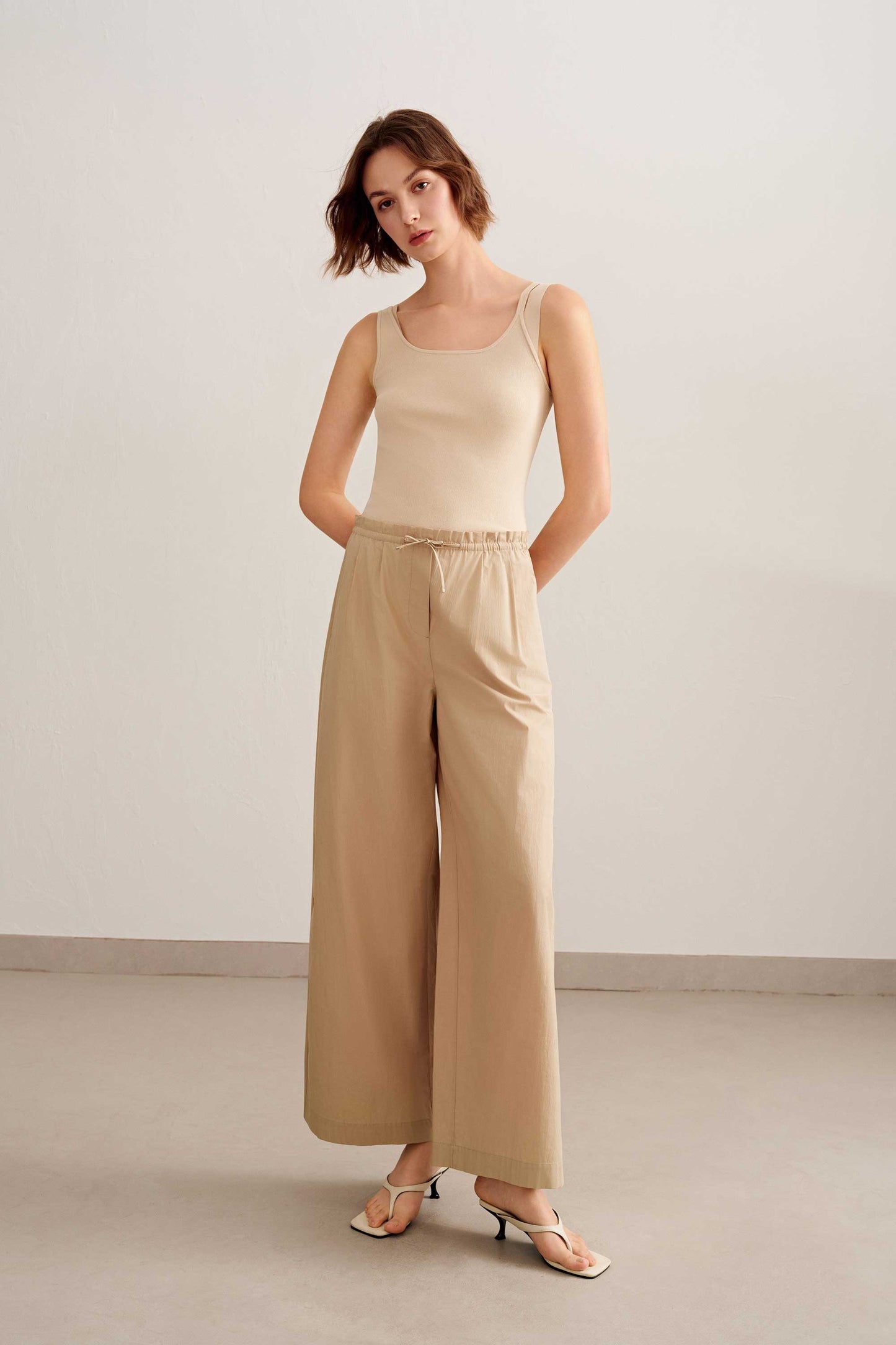 a short brown hair woman wearing a beige tank and a light brown pants white sandal. 
