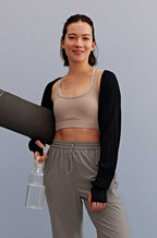a woman holding a water bottle and yoga mat walking, with a light grey sports bra, black shrug and grey pants. 