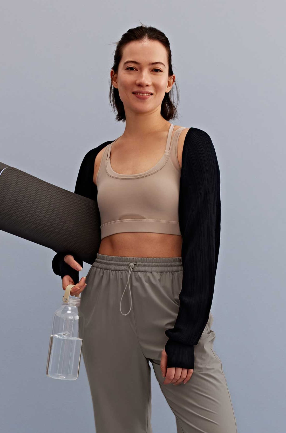 a woman holding a water bottle and yoga mat walking, with a light grey sports bra, black shrug and grey pants. 