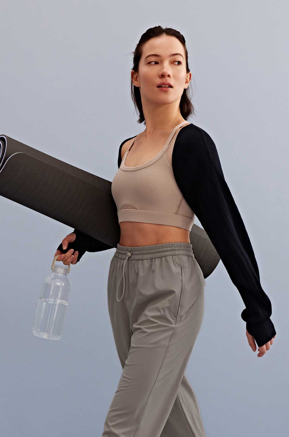 a woman holding a water bottle and yoga mat walking, with a light grey sports bra, black shrug and grey pants. 