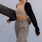 a woman holding a water bottle and yoga mat walking, with a light grey sports bra, black shrug and grey pants. 