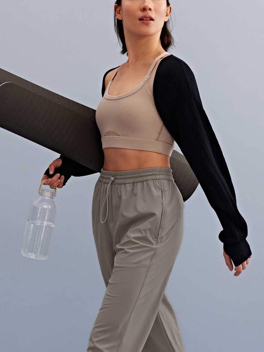 a woman holding a water bottle and yoga mat walking, with a light grey sports bra, black shrug and grey pants. 