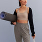 a woman holding a water bottle and yoga mat walking, with a light grey sports bra, black shrug and grey pants. 