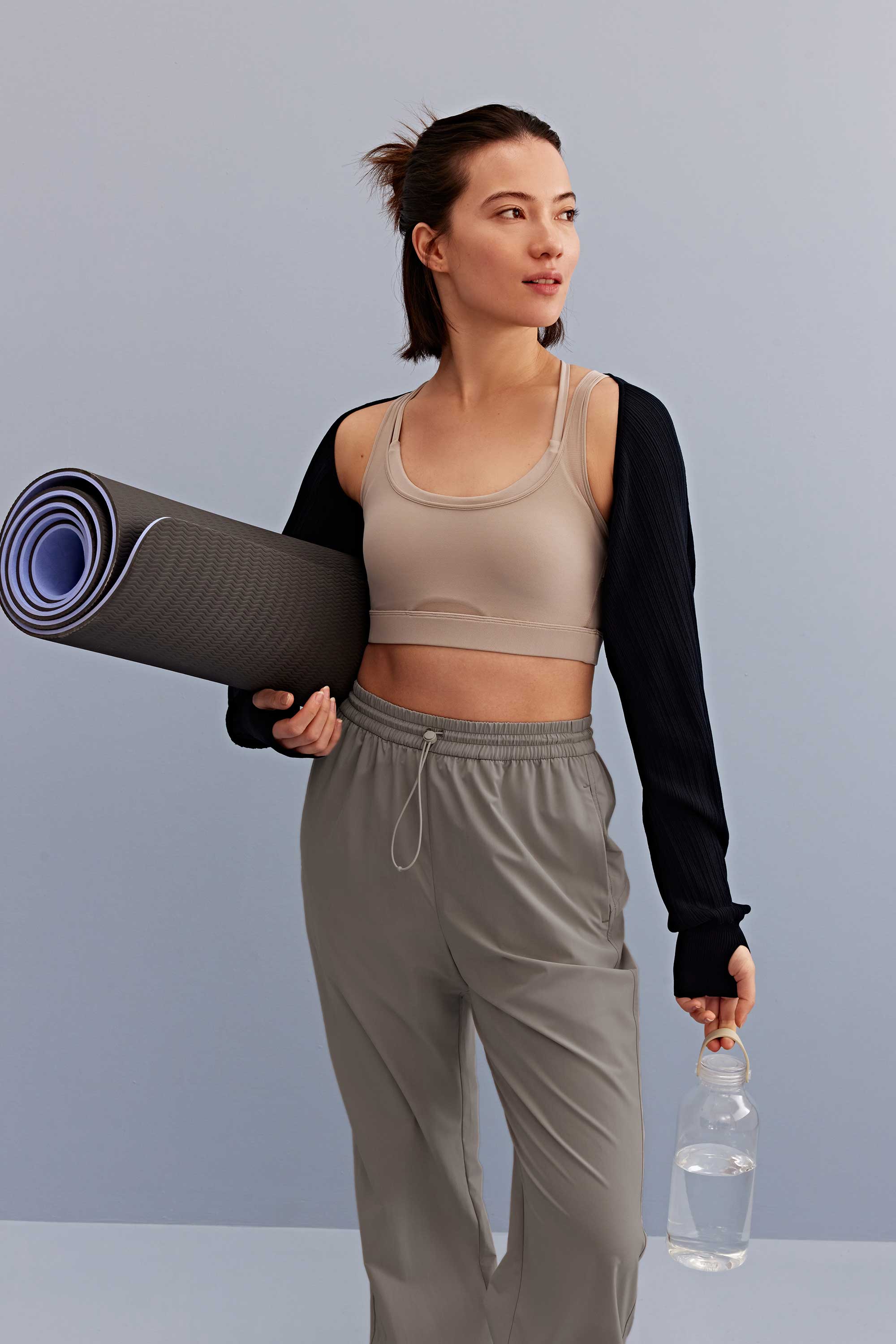 a woman holding a water bottle and yoga mat walking, with a light grey sports bra, black shrug and grey pants. 