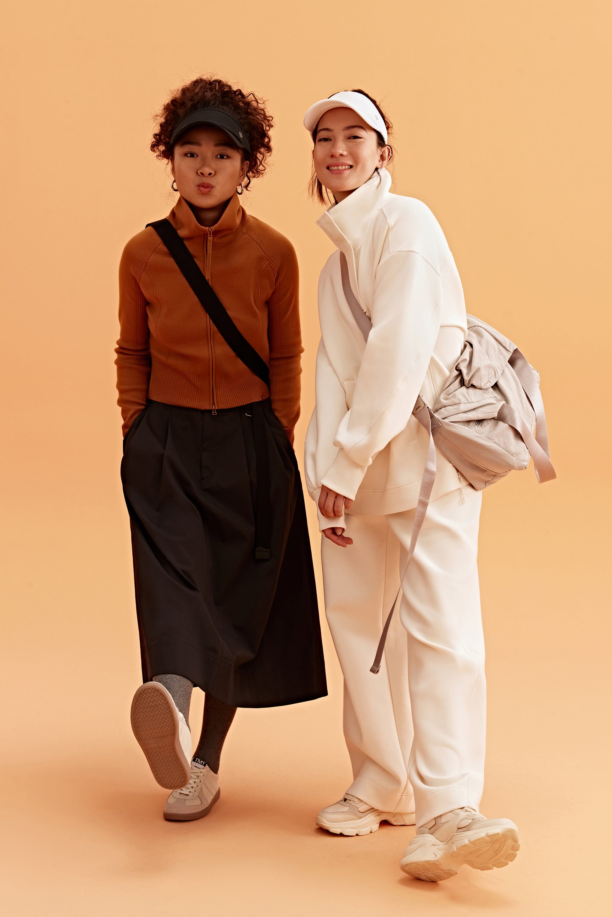 two woman in the photo. the left woman wearing a black visor, brown sweatshirt brown skirt and black bag. the right woman wearing a white visor and white half zip sweatshirt and white pants. pair with a light grey bag