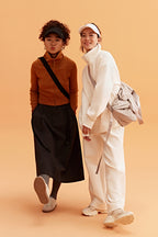 two woman in the photo. the left woman wearing a black visor, brown sweatshirt brown skirt and black bag. the right woman wearing a white visor and white half zip sweatshirt and white pants. pair with a light grey bag