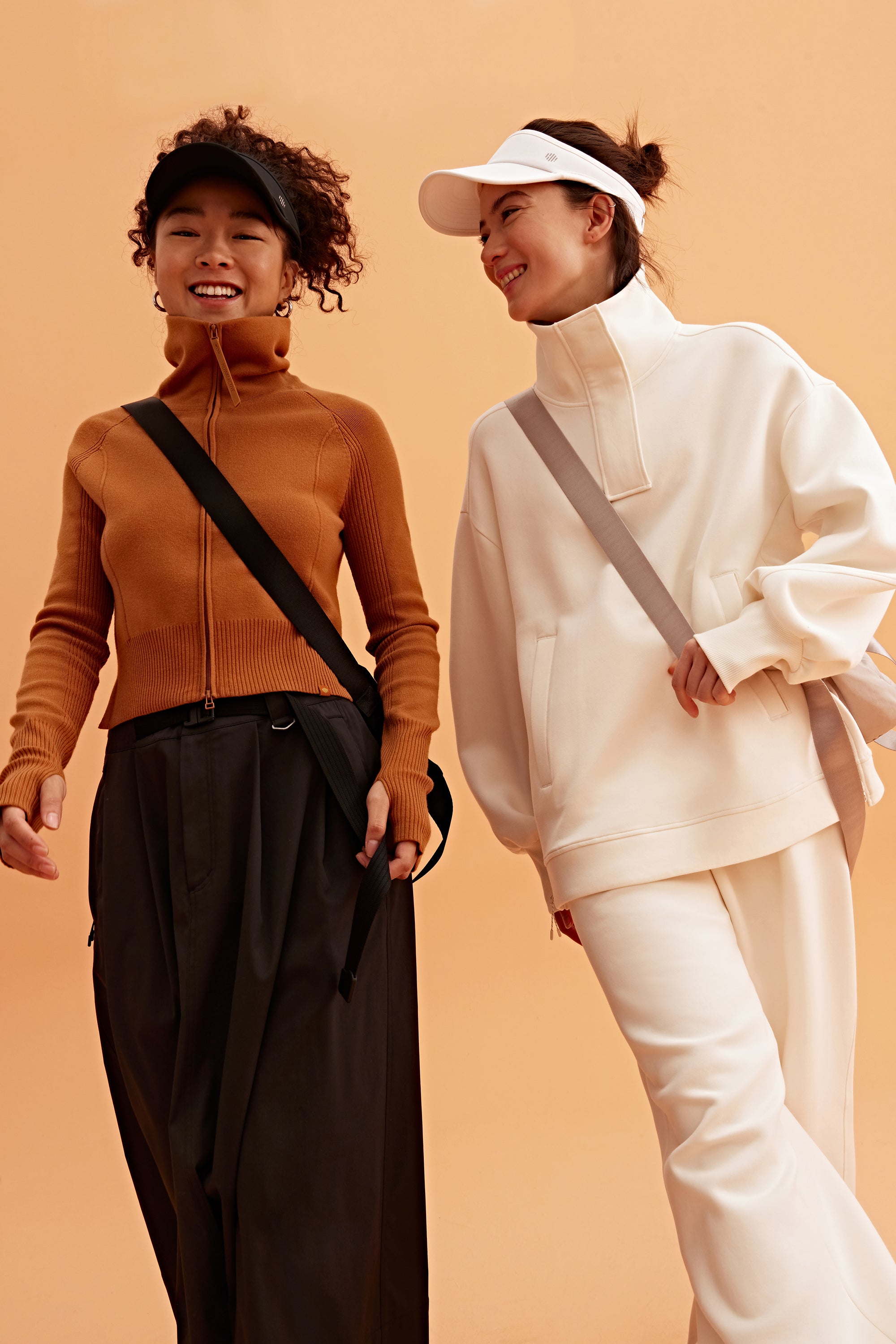 two woman in the photo. the left woman wearing a black visor, brown sweatshirt brown skirt and black bag. the right woman wearing a white visor and white half zip sweatshirt and white pants. pair with a light grey bag