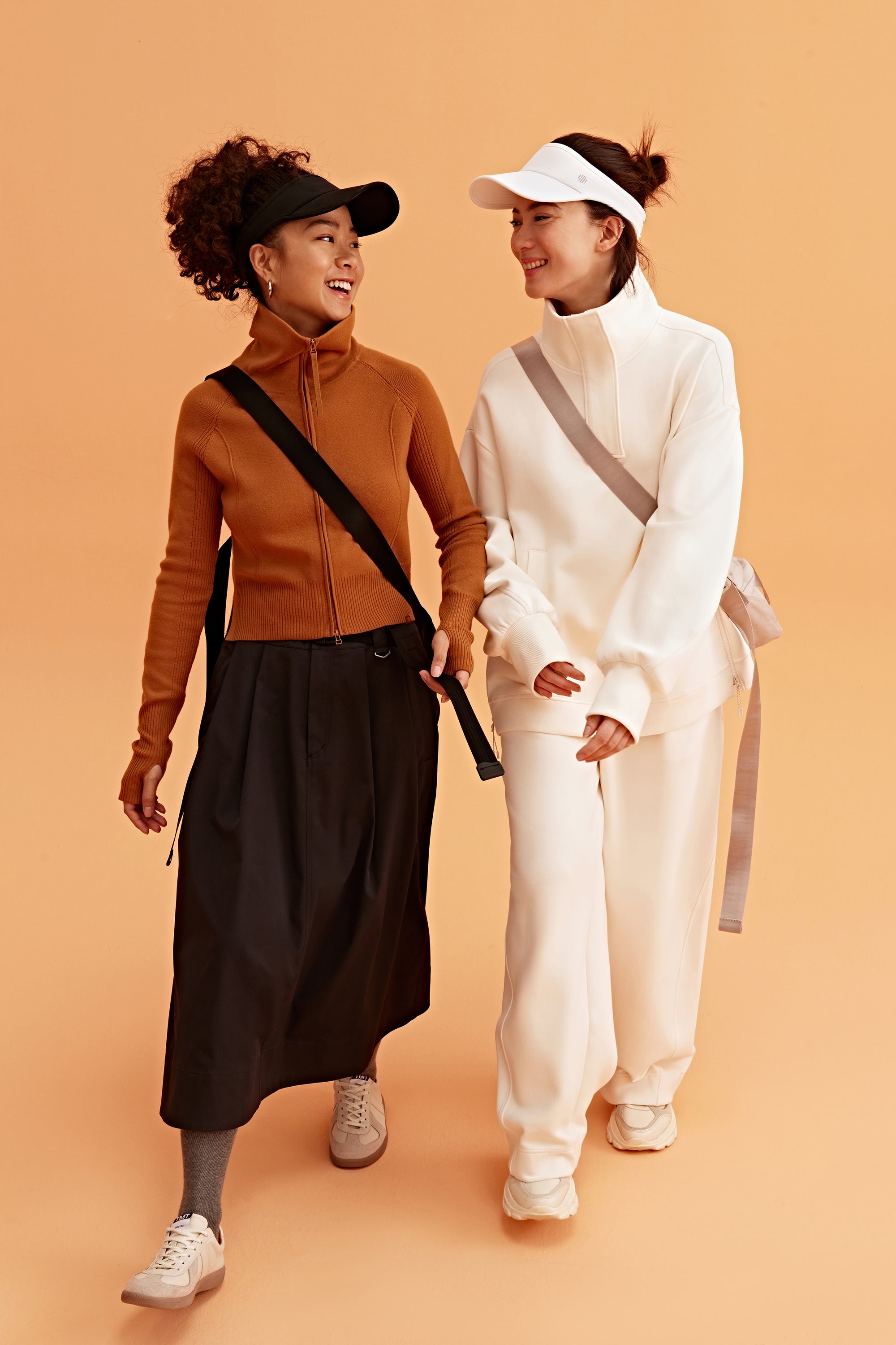 two woman in the photo. the left woman wearing a black visor, brown sweatshirt brown skirt and black bag. the right woman wearing a white visor and white half zip sweatshirt and white pants. pair with a light grey bag