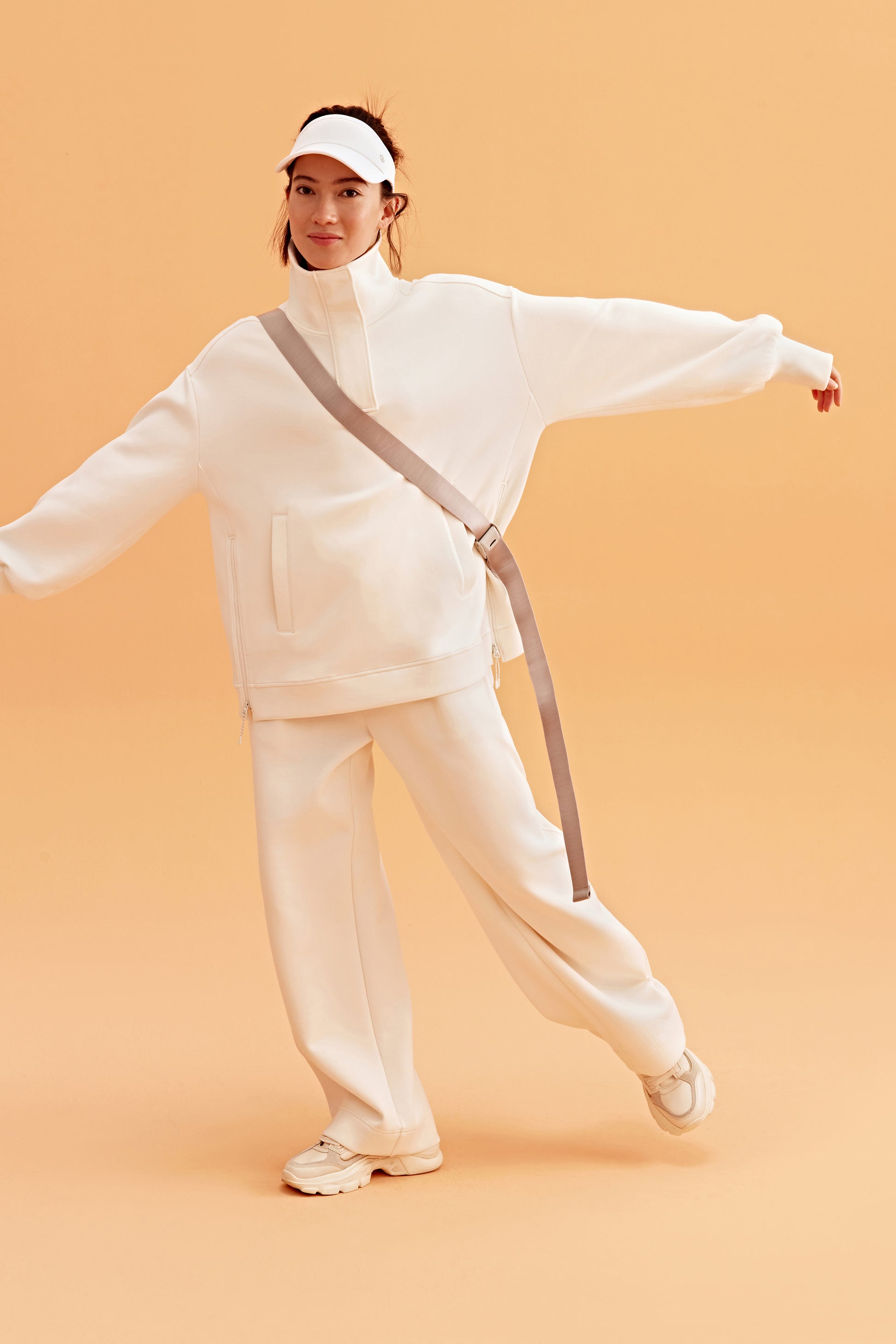 woman wearing a white visor and white half zip sweatshirt and white pants. pair with a white sneakers open her arms