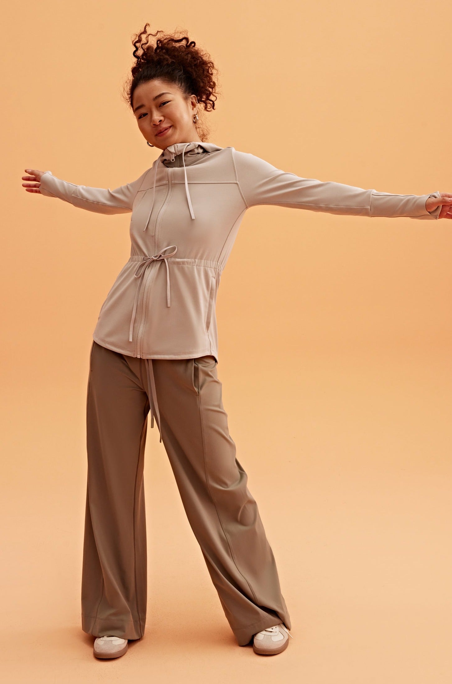 a woman wearing a light brown hooded jacket and brown pants open her arms