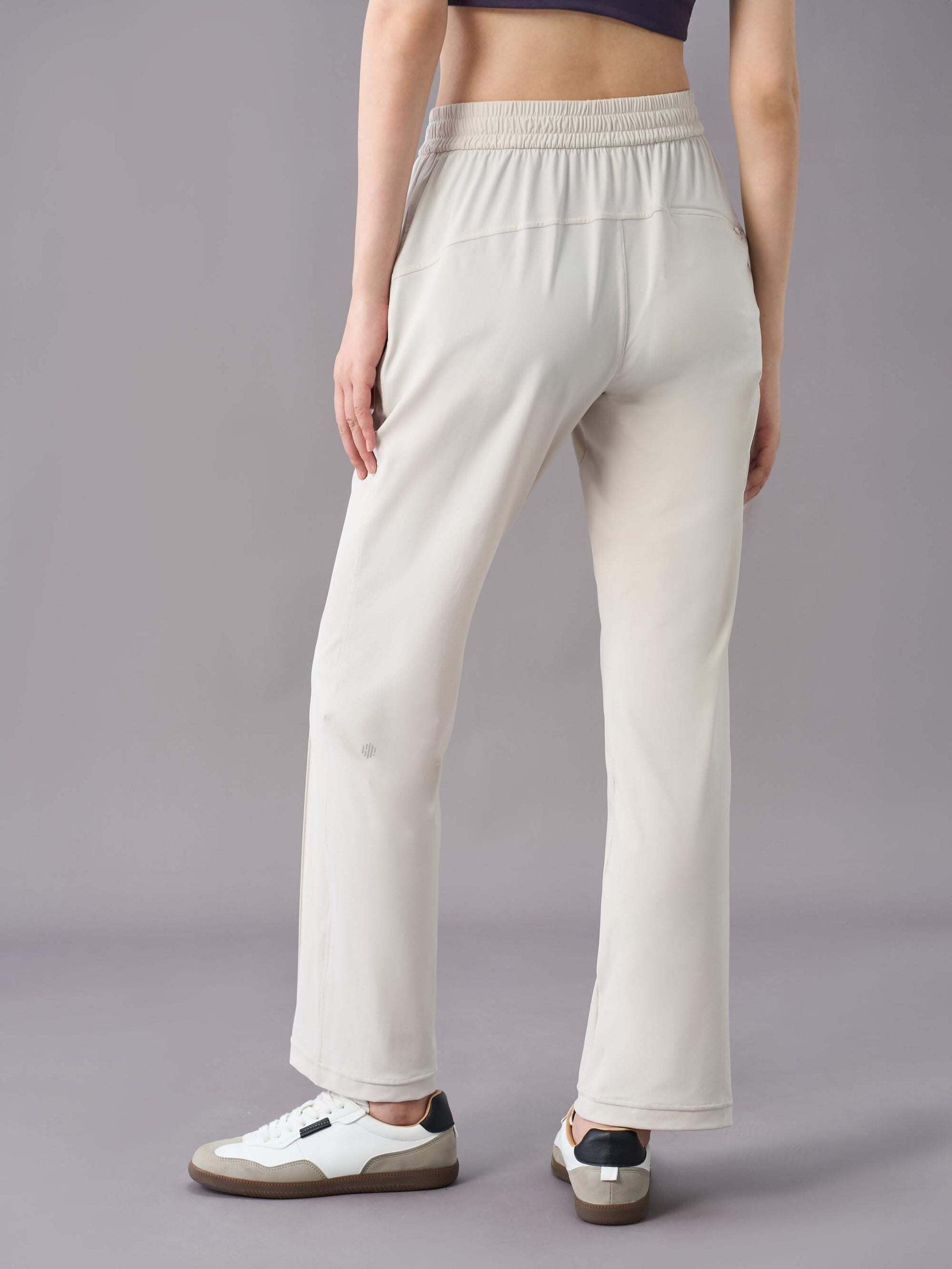 A back view of a model wearing off-white straight-leg pants, paired with white sneakers. The pants feature a relaxed fit with a drawstring waistband, side pockets, and subtle seam details down the front.