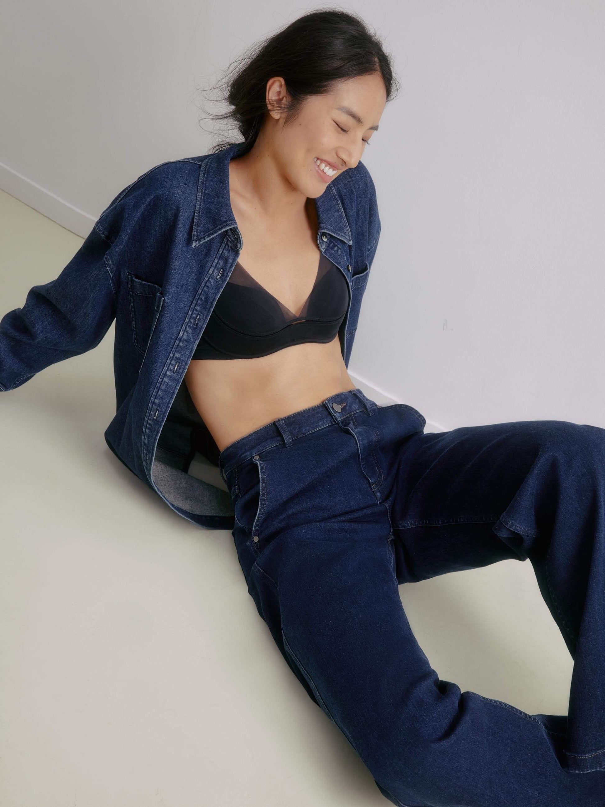 Model wearing a black 3/4 cup bra paired with dark denim jeans and an open denim jacket, sitting on the floor.