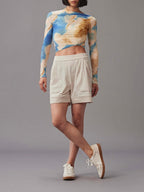 A model wearing a vibrant long-sleeve top, paired with mid-waist light beige shorts. Standing against a grey background. 