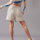 A back view of a model wearing a vibrant long-sleeve top, paired with mid-waist light beige shorts. Standing against a grey background. 