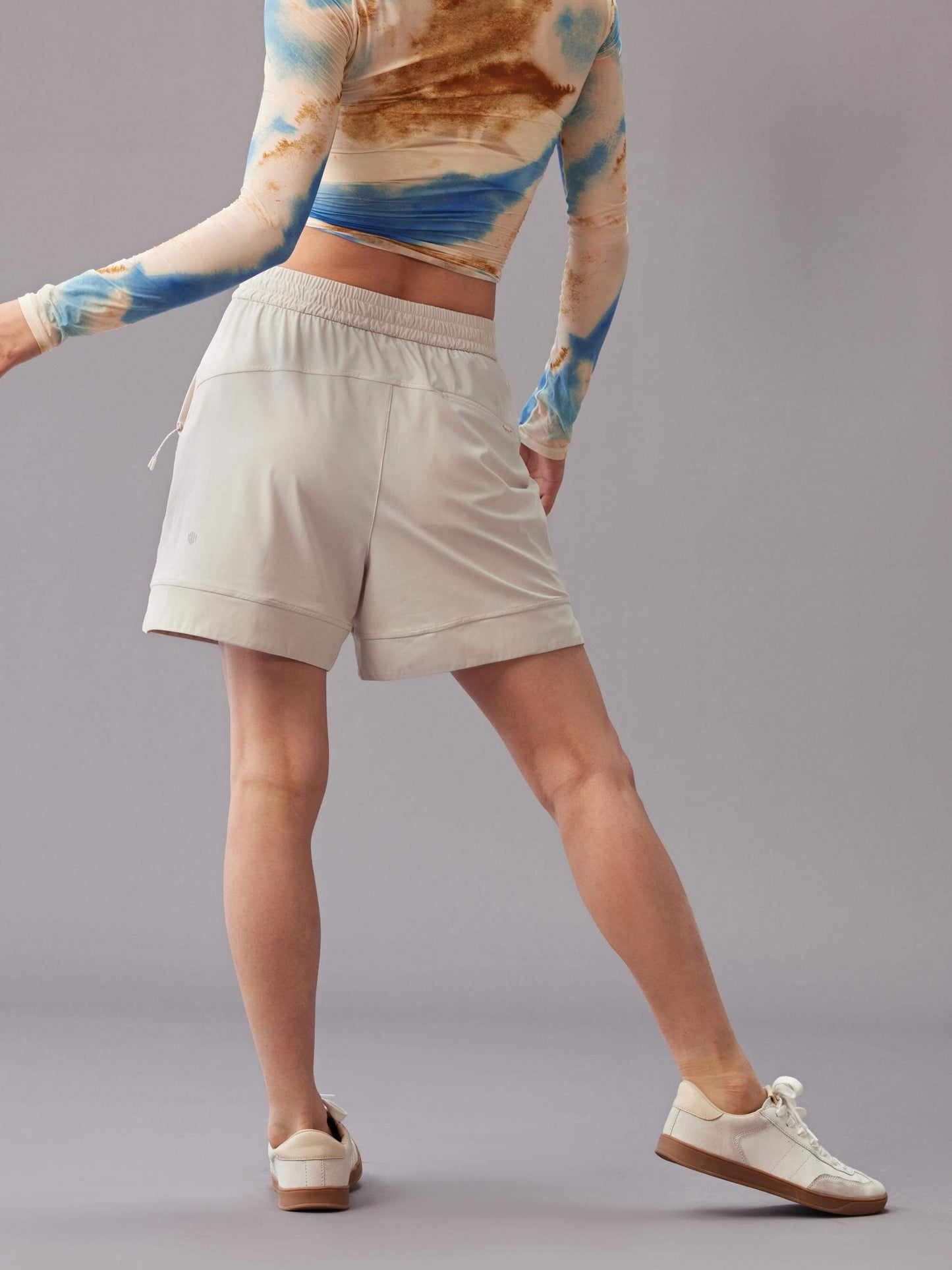 A back view of a model wearing a vibrant long-sleeve top, paired with mid-waist light beige shorts. Standing against a grey background. 