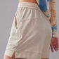 A close-up side view of a model wearing light beige mid-waist shorts paired with a vibrant tie-dye long-sleeve top. The shorts feature an elastic waistband, zippered side pockets.