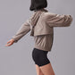 A back view of a model wearing a taupe zip-up sunblock jacket, paired with black high-waisted shorts.