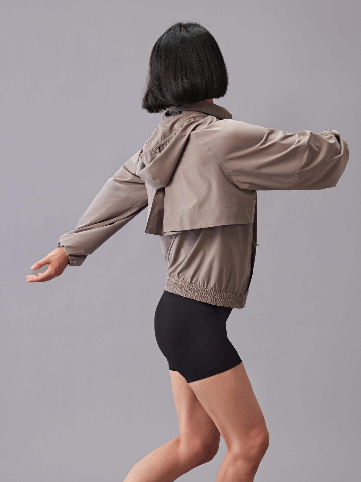 A back view of a model wearing a taupe zip-up sunblock jacket, paired with black high-waisted shorts.